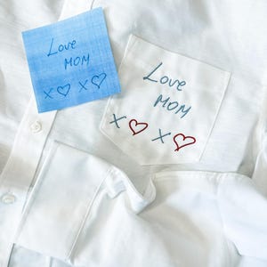 Puede incluir: Una camisa blanca de botones con un bolsillo en el pecho bordado con el texto azul Love MOM y símbolos de X y corazones en rojo y azul. Una nota cuadrada azul con el mismo texto manuscrito y símbolos se coloca sobre la tela.