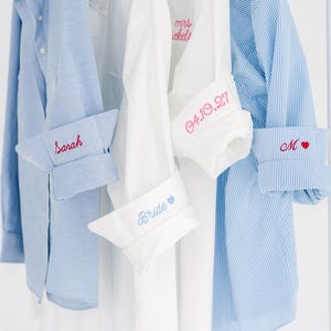 May include: A collection of long-sleeved button-down shirts in white and light blue, including solid and pinstriped patterns. Each shirt features personalized embroidery on the cuffs, displaying names, dates, and symbols like hearts in various colors.