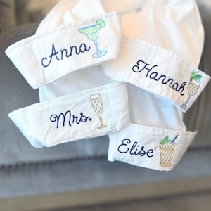 May include: Four white fabric cuffs featuring navy blue embroidered names and colorful cocktail illustrations. The names Anna, Hannah, Mrs., and Elise are stitched alongside a margarita, a martini glass, a champagne flute, and a highball glass.