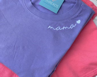 Puede incluir: Una camiseta de cuello redondo color lavanda con el texto blanco bordado "mama" seguido de un pequeño corazón blanco en la parte superior izquierda del pecho. Una etiqueta cuadrada azul con el texto "7 THREADS" está unida al cuello. Se ve una camiseta rosa.