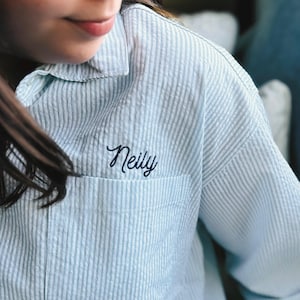 May include: Light blue and white striped pajama shirt with a pocket. The name "Neilly" is embroidered in a cursive font on the pocket. The shirt has a classic collar and button-down front.