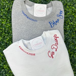 Op de afbeelding: Twee sweatshirts met geborduurde tekst. Het bovenste sweatshirt is grijs met blauwe borduurwerk dat "Blue Sox" zegt. Het onderste sweatshirt is wit met rode borduurwerk dat "Go Dawgs" zegt.