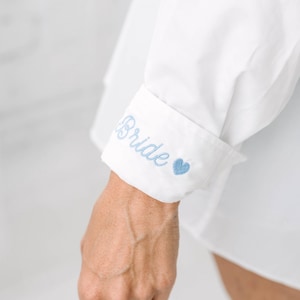 May include: A white long-sleeved shirt featuring a folded cuff with the word Bride embroidered in light blue script lettering, followed by a small light blue embroidered heart symbol on the outer edge of the fabric cuff.