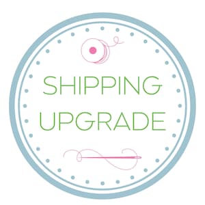 May include: A circular graphic with a light blue border and dotted accents. The words "SHIPPING UPGRADE" are in green, centered within the circle. A pink spool of thread and needle are at the top and bottom.
