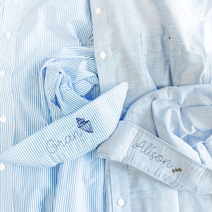 Könnte beinhalten: Zwei hellblaue Hemden mit Knöpfen. Eines ist gestreift, das andere uni. Das Manschetten der gestreiften Hemden hat den Namen "Oran" in Blau gestickt, zusammen mit einem Muscheldesign. Die Manschette des einfarbigen Hemdes hat den Namen "Alison" in Blau gestickt.