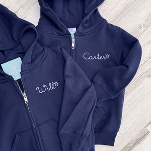 May include: Two navy blue zip-up hoodies with white embroidered names and a star. The hoodie on the left says "Will" and the hoodie on the right says "Carter".