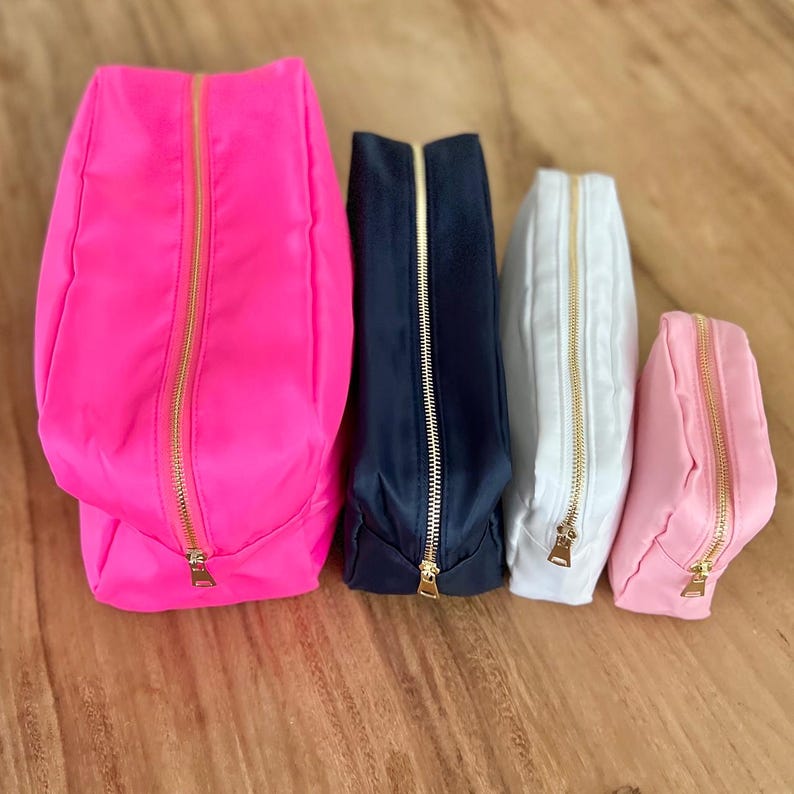 May include: Four zippered cosmetic bags in varying sizes and colors. The bags are arranged from largest to smallest, with colors including hot pink, navy blue, white, and light pink. Each bag has a gold-colored zipper.