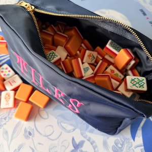 May include: Navy blue zippered pouch with the word "ILLES" embroidered in pink. The pouch is filled with orange and white Mahjong tiles. The gold zipper is open, revealing the tiles inside. Some tiles are scattered outside the pouch.