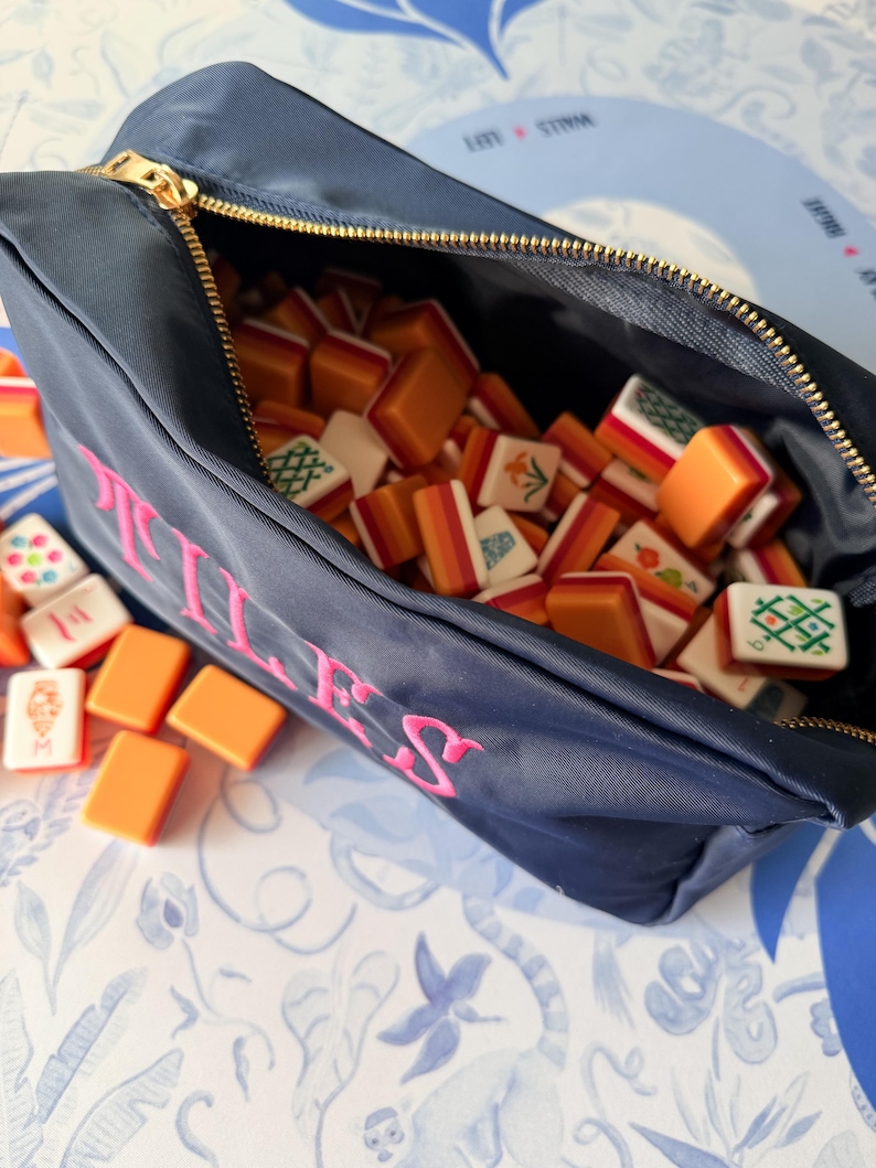 May include: Navy blue zippered pouch with the word "ILLES" embroidered in pink. The pouch is filled with orange and white Mahjong tiles. The gold zipper is open, revealing the tiles inside. Some tiles are scattered outside the pouch.