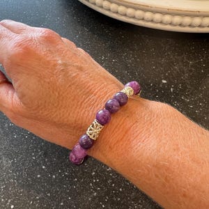 May include: A purple gemstone bracelet with gold accents. The bracelet is made of round beads and has a simple design.