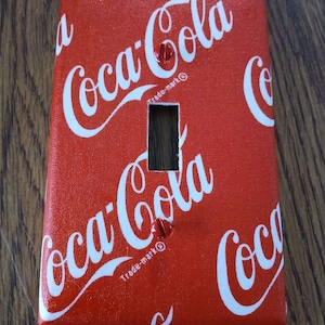 May include: Red light switch cover with a Coca-Cola pattern. The cover has a white background with the Coca-Cola logo repeated in white lettering.