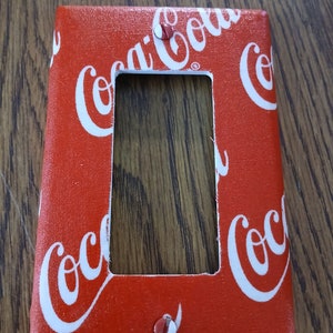 May include: Red light switch cover with a Coca-Cola pattern. The cover has a white rectangular opening for the switch.