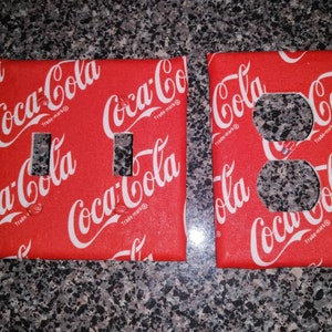 May include: Two red light switch covers with a Coca-Cola pattern. The covers have two light switches and one outlet.