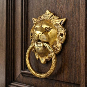 May include: A gold-colored lion head door knocker with a ring handle, mounted on a dark brown wooden door. The lion's face has detailed features, and the ring handle is textured.