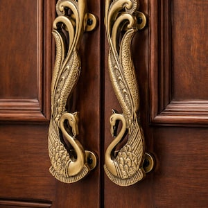 Puede incluir: Manijas de puerta de latón dorado ornamentadas con un diseño de cisne. Las manijas están montadas en una puerta de madera marrón oscuro. Las alas y el cuello del cisne están detallados, y las manijas tienen un diseño decorativo y fluido.