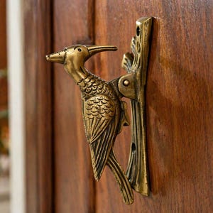 May include: A brass door knocker in the shape of a bird, mounted on a wooden door. The bird has detailed feather and wing patterns. The knocker is a decorative element for a front door.