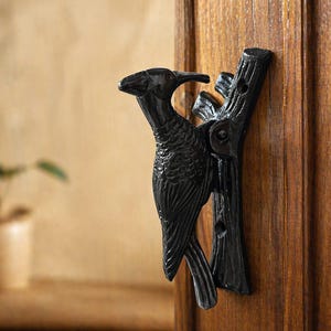 Op de afbeelding: Zwarte deurklopper van gietijzer in de vorm van een vogel op een tak. De vogel heeft gedetailleerde veren en een lange snavel. De deurklopper is gemonteerd op een houten deur.