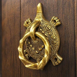 Op de afbeelding: Een gedetailleerde, antieke messing deurklopper in de vorm van een schildpad, gemonteerd op een donkere houten deur. De klopper heeft een sierlijk ontwerp met een ring om op te kloppen. Het messing heeft een warme, gouden tint.