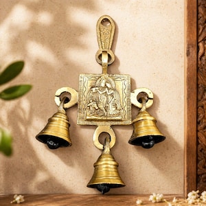 Könnte beinhalten: Ein Wandbehang aus Messing mit drei Glocken und einem zentralen Quadrat mit einer eingravierten religiösen Szene. Die Glocken sind an einem dekorativen Rahmen aufgehängt und schaffen eine traditionelle Ästhetik. Das Stück ist aus Messing.