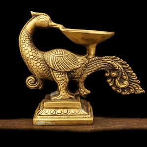 May include: A gold-coloured peacock-shaped oil lamp, with detailed feather and body patterns. The lamp has a small bowl held by the peacock's beak. It stands on a square base. The background is black.