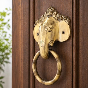May include: A brass elephant-shaped door knocker with a ring handle. The knocker is mounted on a dark brown wooden door. The elephant head features detailed carvings and a decorative pattern on the trunk.