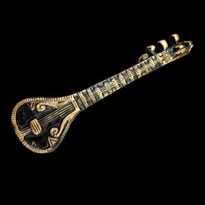 May include: A detailed, gold-coloured miniature sitar against a black background. The instrument features intricate carvings and a decorative design on the body. The sitar's neck is long and slender, with tuning pegs at the top.