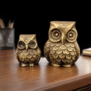 Op de afbeelding: Twee messing uilbeeldjes, één groot en één klein, staan naast elkaar op een houten oppervlak. De uilen hebben gedetailleerde veermotieven en grote, ronde ogen. De donkere achtergrond benadrukt de gouden kleur van de sculpturen.