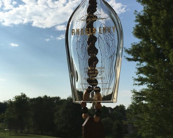 May include: A clear glass wind chime made from a repurposed bottle, featuring the "Angels Envy" logo and angel wing design. The chime has a chain, wooden beads, and a dark brown tag. The background shows a blue sky and green trees.