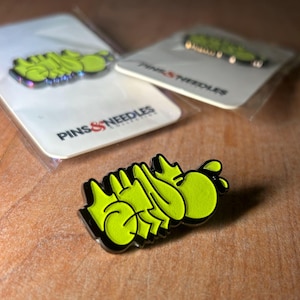 May include: Close-up of three enamel pins with a graffiti-style design in neon green and black. The pins are on white backing cards with the text "PINS & NEEDLES" and are packaged in clear plastic.