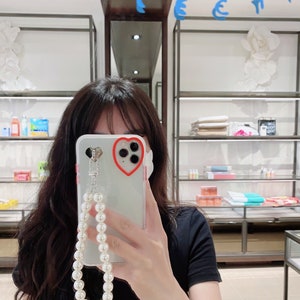 May include: A clear phone case with a red heart cutout around the camera lens. The case has a pearl chain attached to it.