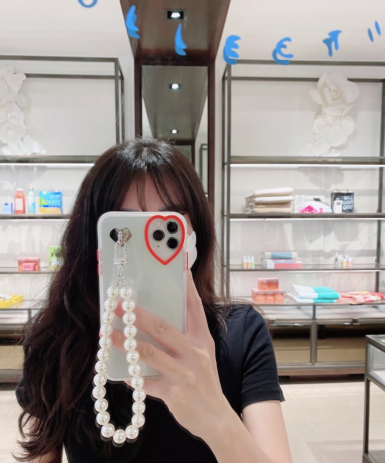 May include: A clear phone case with a red heart cutout around the camera lens. The case has a pearl chain attached to it.