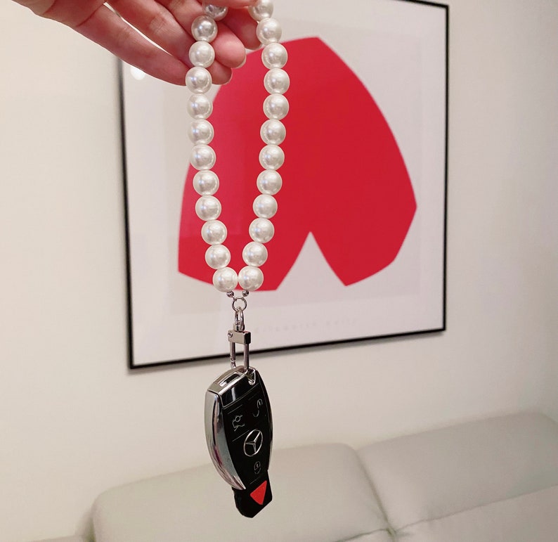 May include: A white pearl necklace with a silver key chain attached to the end. The key chain has a black car key with silver buttons.