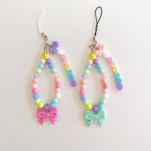 May include: Two colorful beaded phone charms with a smiley face charm and a bow at the bottom. One charm has a pink bow and the other has a light blue bow.
