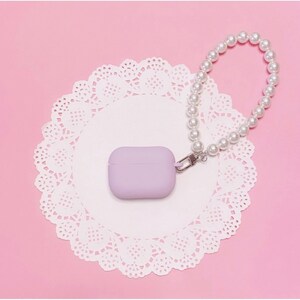 May include: A lavender silicone case for AirPods with a pearl chain attached. The case and chain are on a white doily on a pink background.