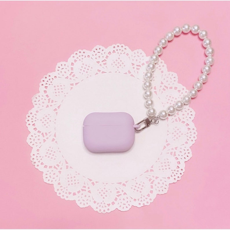 May include: A lavender silicone case for AirPods with a pearl chain attached. The case and chain are on a white doily on a pink background.