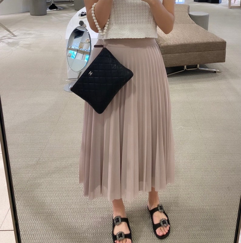 May include: A black quilted clutch bag with a pearl chain strap. The bag is being held by a person wearing a white top and a beige pleated skirt.