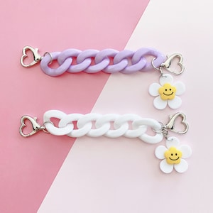 May include: Two plastic chain phone charms with a smiley face daisy charm attached to each. One chain is purple and the other is white.