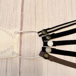 May include: Five different mask extenders with various colors and patterns. The extenders are made of fabric and have a button on each end. The buttons are black, white, gray, and brown.