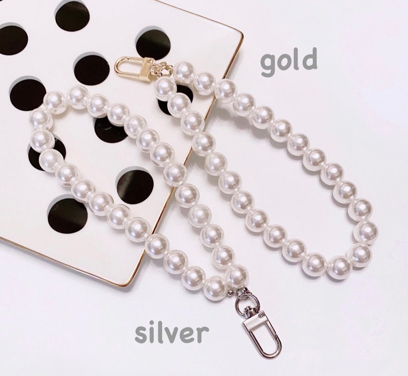 May include: A white pearl chain bag strap with gold and silver clasps. The strap is on a white surface with black polka dots. The words "gold" and "silver" are written below the strap.