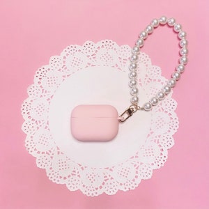May include: A pink Airpods case with a pearl chain attached. The case is sitting on a white doily on a pink background.