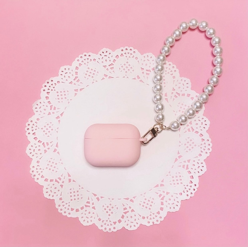 May include: A pink Airpods case with a pearl chain attached. The case is sitting on a white doily on a pink background.