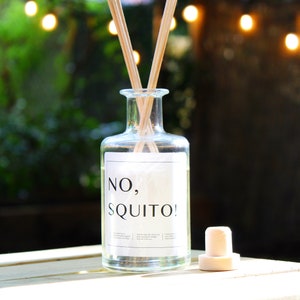 May include: A clear glass bottle with a white label that reads "NO, SQUITO!" and a cork stopper. The bottle is filled with a clear liquid and has wooden sticks inserted into it. The bottle is sitting on a wooden surface with a blurred background of green foliage and string lights.
