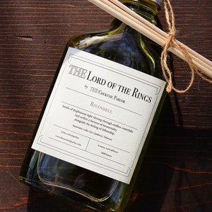 May include: A clear glass bottle with a white label that reads "The Lord of the Rings" by "The Cocktail Parlor". The label also says "Rivendell" and describes the scent as "Smells of dappled light shining through endless waterfalls laid within a forest of immortality alongside the feeling of fellowship". The bottle is sitting on a wooden surface with three wooden sticks tied together with twine.