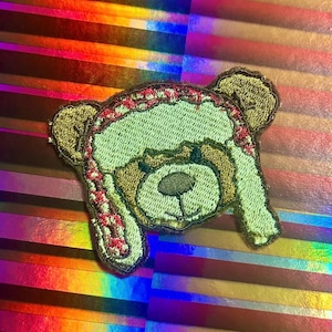 May include: An embroidered patch featuring a brown bear head wearing a light green winter hat with a red and white patterned trim. The bear has dark brown ears, a light green muzzle, and a dark brown nose with stitched facial details.