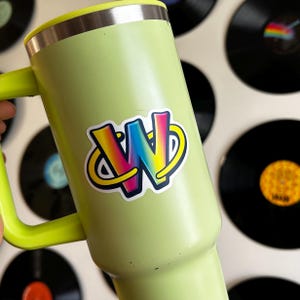 Puede incluir: Un termo de viaje verde claro con borde plateado y asa verde brillante. Una pegatina colorida con las letras "WN" en colores arcoíris está adherida al termo. El fondo presenta varios discos de vinilo negros.