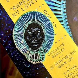 Pode incluir: Patch bordado com um rosto negro e um design de halo azul e verde-azulado. O patch está sobre um fundo amarelo com texto que inclui "AWAKE MY LOVE" e "EXCLUSIVE BOOKLET". Provavelmente para roupas ou acessórios.