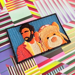 May include: Embroidered patch featuring a man with a beard and a teddy bear. The patch has a blue background and a black border. The man is wearing a red jacket and a white shirt. The teddy bear is brown.