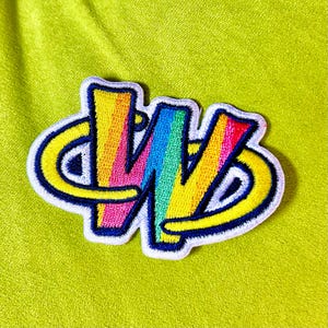 May include: Embroidered patch with a rainbow-coloured 'W' design, outlined in yellow and blue, with a white border. The patch is set against a lime green background. Suitable for customising clothing or accessories. The patch measures approximately 7 cm across.