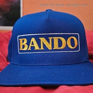 May include: A blue baseball cap with a white and yellow embroidered rectangle that says "BANDO" on it.