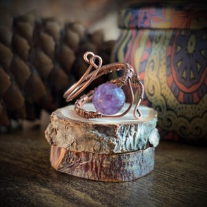 May include: A handcrafted copper wire ring featuring a round, purple amethyst gemstone. The ring is artistically wrapped and sits on a small, rustic wooden display. The background includes a decorative jar and a pine cone.